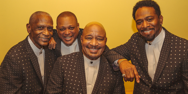 The 4 members of The Stylistics in suits against a yellow background.