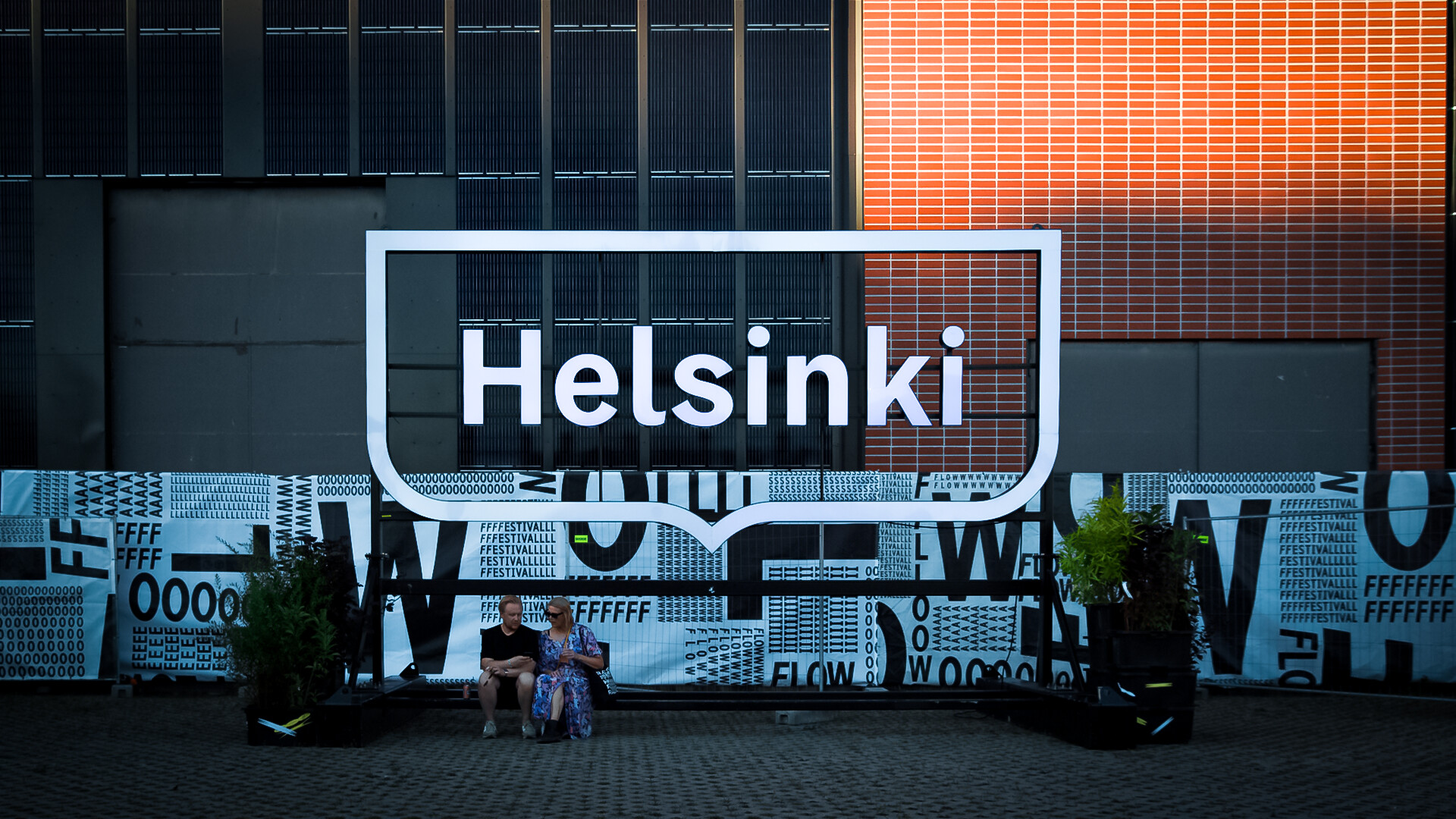 Valokuva. Nuoria henkilöitä istmuassa kadulla. Taustalla Helsinki -logo.
