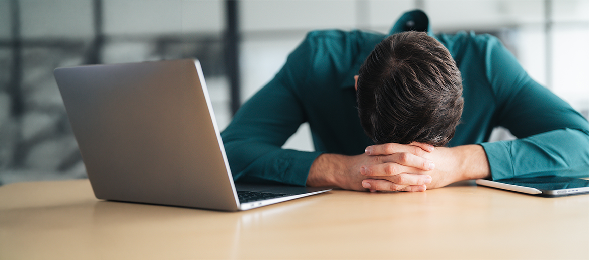 Person mit gesenktem Kopf am Schreibtisch neben Laptop und Smartphone, Büro, Erschöpfung im Arbeitsalltag, Stress, Homeoffice. Fotorealistische Darstellung einer Person empfohlen.