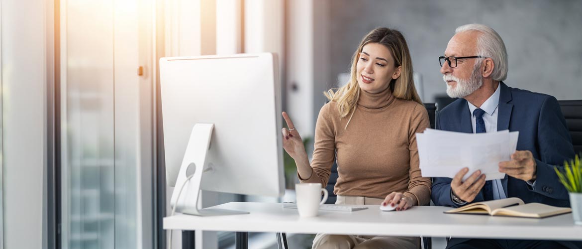 Zwei Personen arbeiten gemeinsam im modernen Büro am Schreibtisch, mit Desktop‑Computer, Dokumenten und Notizbuch, Business‑Meeting, Teamarbeit, Büroalltag. Fotorealistische Abbildung verwenden.