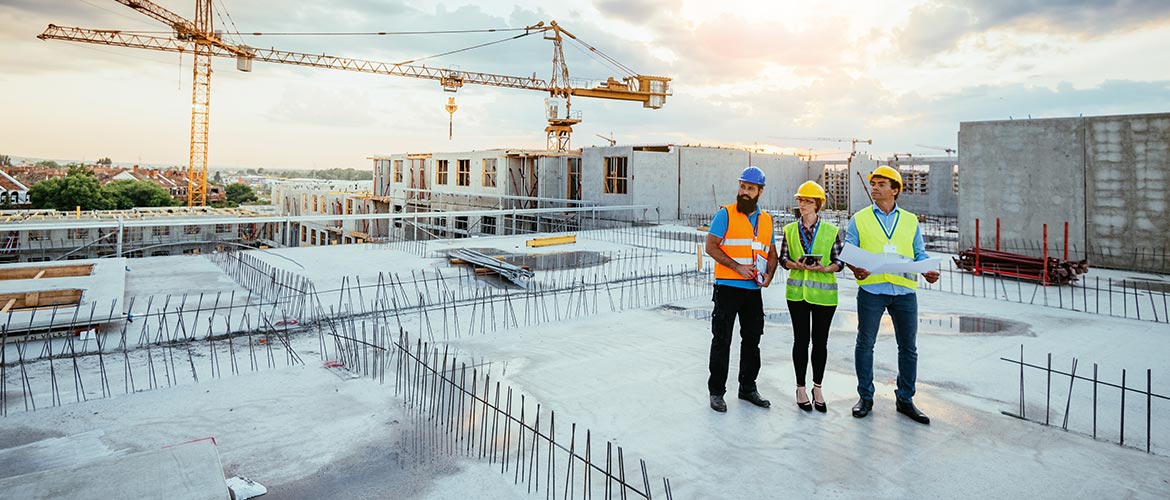 Baustelle mit drei Personen in Sicherheitskleidung auf einer Rohbaufläche, umgeben von Kränen und Betonstrukturen. Keywords: Baustelle, Rohbau, Bauplanung, Arbeitssicherheit, Konstruktion.