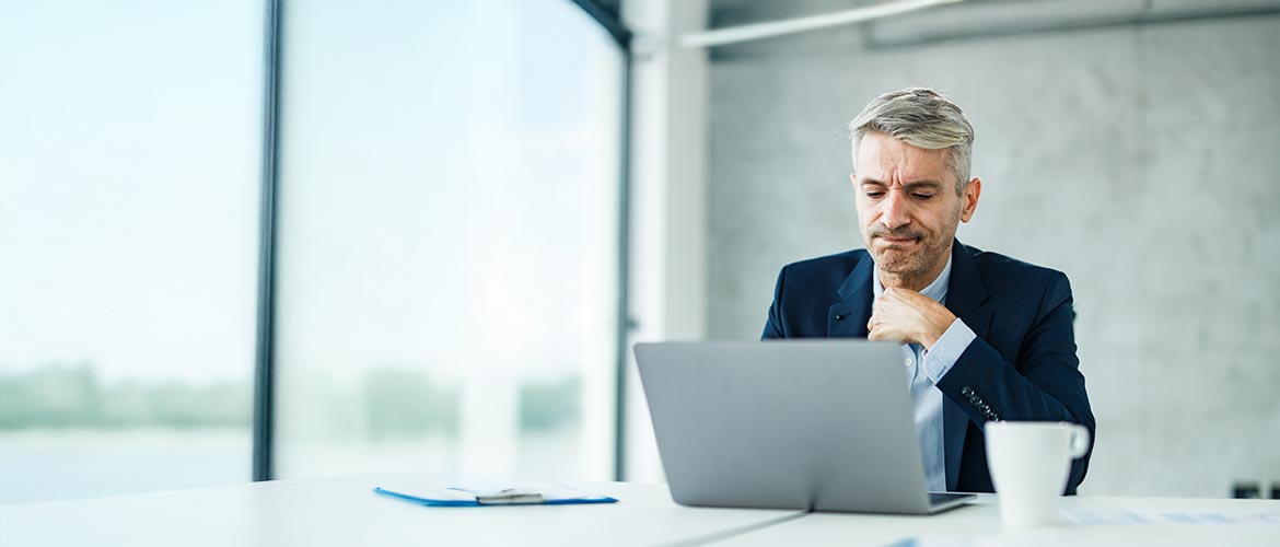 Moderner Büroarbeitsplatz mit einer Person im Business‑Anzug am Laptop, hellem Konferenzraum, Unterlagen und Kaffeetasse.