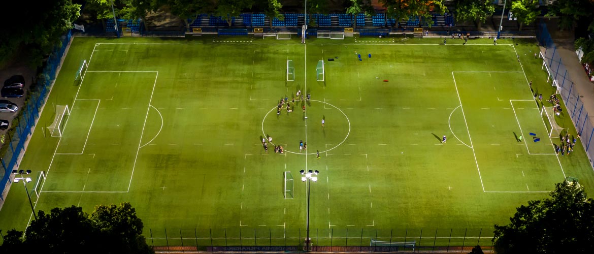 Beleuchteter Fußballplatz bei Nacht mit Trainingsgruppen, grünem Kunstrasen, Toren und Symmetrie aus der Vogelperspektive. Ideal für Sport, Training, Fußballplatz, Nachtaufnahme.