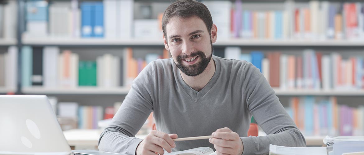 Mann sitzt mit einem Laptop vor einem offenen Buch vor ihm und lächelt vor einem Bücherregal im Hintergrund