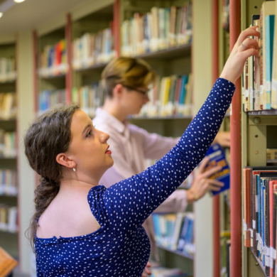 A student reaching for a library book on a shelf