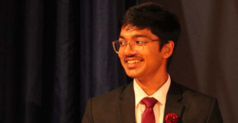 Saswat is wearing a red tie, black suit and glasses. Behind him is a black curtain.