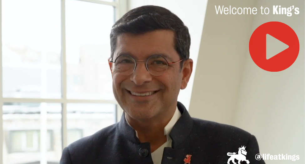 Professor Shitij Kapur stands in front of a window and smiles at the camera. He is wearing round glasses, a white shirt and dark coat with a Reggie the Lion pin on the lapel