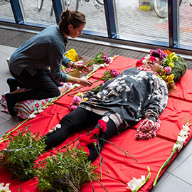Henkilö makaa punaisella pajalla ja toinen henkilö peitää häntä kukilla sekä kasveilla.