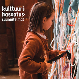 Lapsi seisoo kangassuikaleista tehdyn seinän edessä ja tunnustelee seinästä roikkuvia suikaleita. Taustalla on teksti: kulttuurikasvatussuunnitelmat.