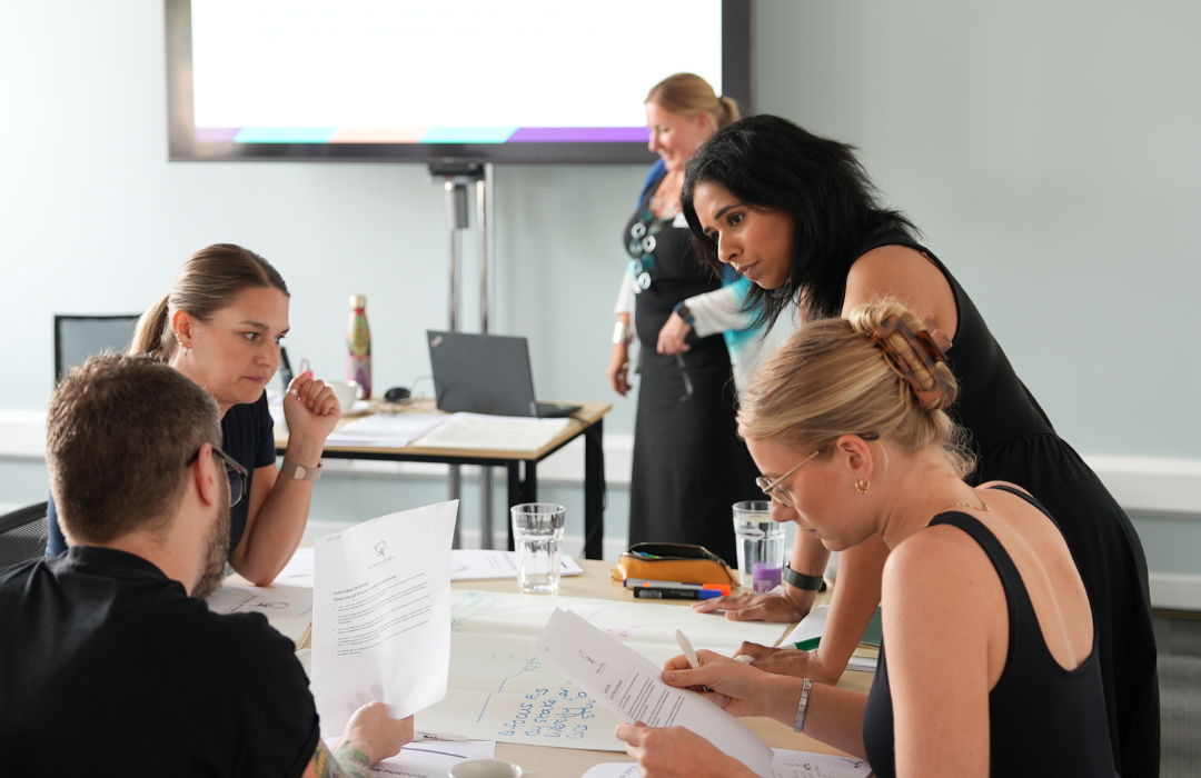 A group of adults in an office space with a projector screen. They are sitting, or standing, around a table and working on a project together involving large sheets of paper and ideas written on the paper.