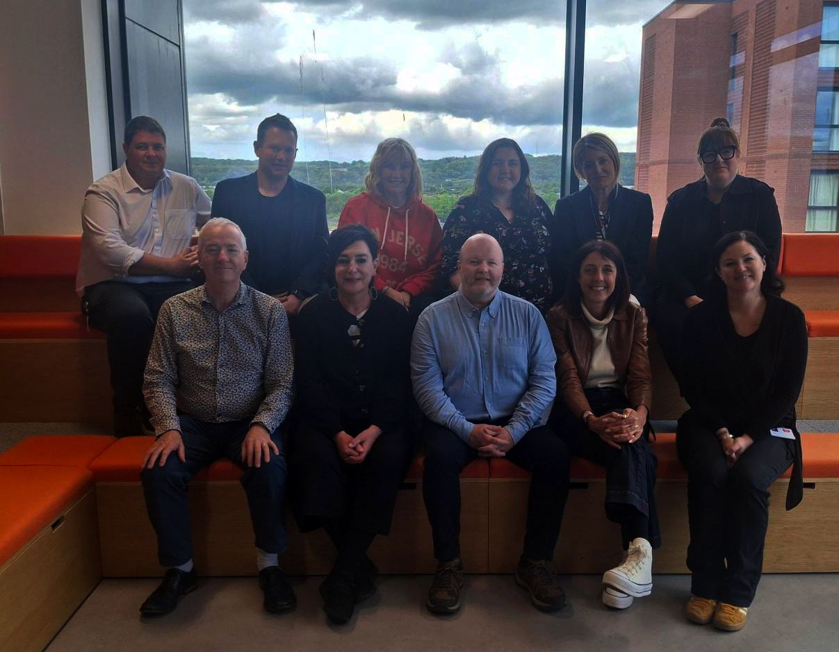 Group photo of ten adults seated and standing in a modern indoor space with tiered wooden seating and large windows behind them. The group is smiling toward the camera, with cloudy skies and buildings visible through the windows in the background. Some people are dressed casually while others wear business attire.