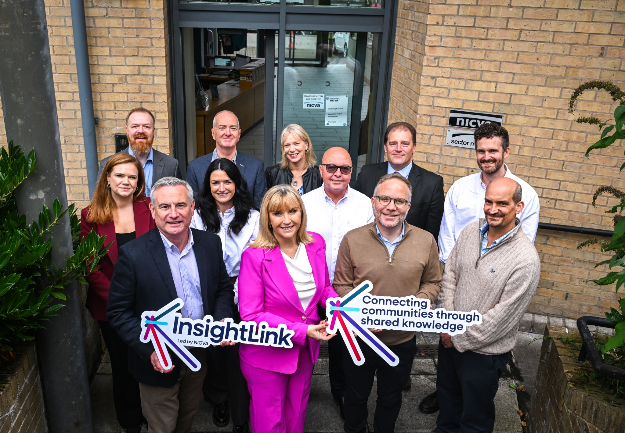 A group poses outside NICVA’s offices, holding signs that read “InsightLink” and “Connecting communities through shared knowledge.”