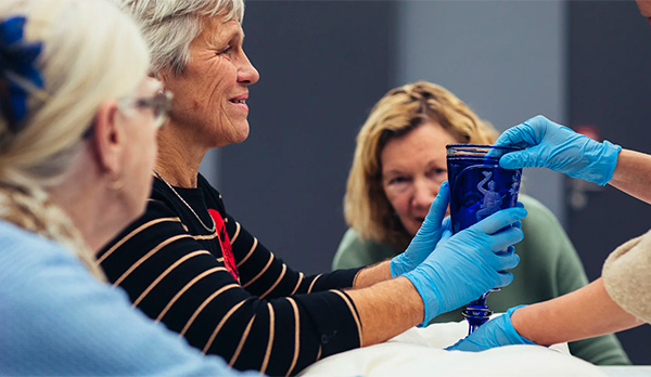 Een groep mensen neemt deel aan een interactieve museumervaring. Een vrouw met blauwe handschoenen reikt een prachtige blauwe glazen bokaal aan, terwijl anderen aandachtig toekijken.