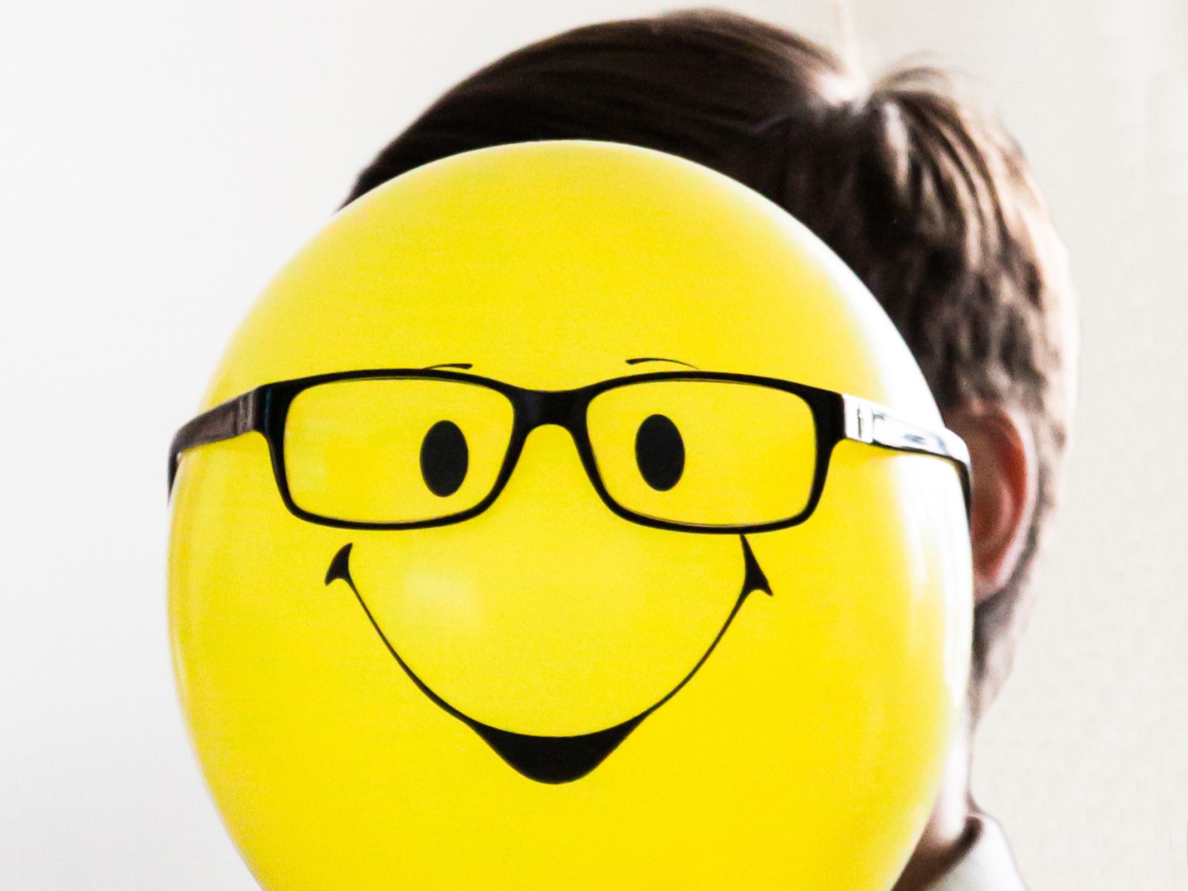 A persons head blocked by a yellow balloon that is smiling and wearing glasses