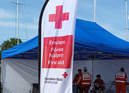 Taustalla teltta ja Punaisen Ristin vapaaehtoisia. Edessä surffi-lippu, jossa lukee Ensiapu eri kielillä.