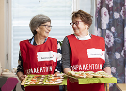 Kaksi Punaisen Ristin vapaaehtoista seisoo rinnakkain katsoen toisiinsa. Käsissään heillä on tarjottimet, joissa on voileipiä. 