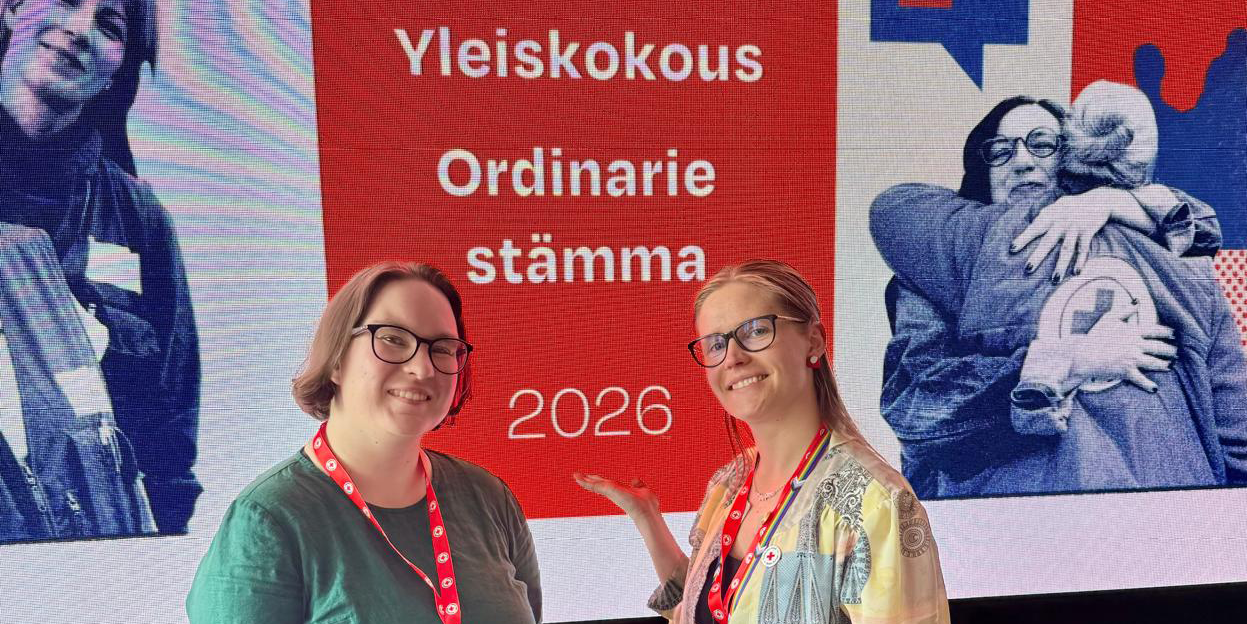 Kaksi hymyilevää henkilöä. Taustalla screeni, jossa teksti Yleiskokous, Ordinarie, Stämma 2026.