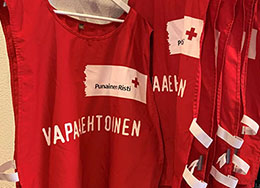 Punaisen Ristin lippuja liehuu sinistä taivasta ja mäntyjä vasten.