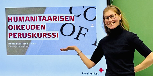 Hymyilevä henkilö näyttää kädellään tekstiä, jossa lukee "Humanitaarisen oikeuden peruskurssi".