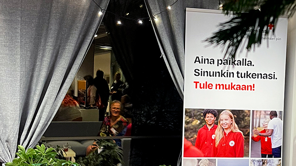 Ikkunasta heijastuu sali, jossa glögikutsujen viettäjiä. Edessä roll-up, jossa teksti "Aina paikalla. Sinunkin tukenasi. Tule mukaan!"