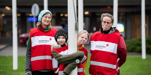 Punaisen Ristin vapaaehtoisia liiveissä, kaksi aikuista ja kaksi lasta. Lapset halaavat lipputankoa.