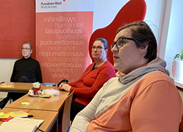 Kolme henkilöä istuu pöytien ääressä. Taustalla näkyy Punaisen Ristin periaate-rolluppia.