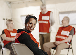 Punaisen Ristin vapaaehtoinen istuu kasvot kohti kameraa. Taustalla kolme muuta vapaaehtoista. 