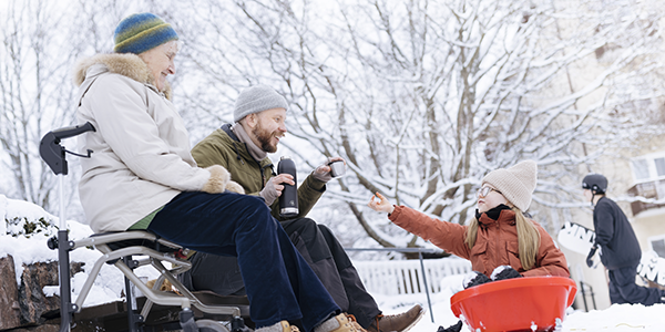 Henkilöitä ulkona talvisessa säässä. Lapsi pulkassa vastaanottamassa juotavaa termoskannusta.