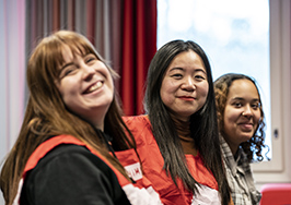 Kolme hymyilevää Punaisen Ristin vapaaehtoista istuu rinnakkain sisätiloissa.