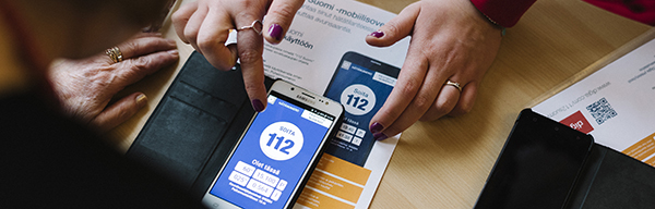 Kännykkä on pöydällä. Kännykässä on avoinna 112 Suomi -sovellus ja henkilön kädet opastamassa sen käyttöä. 